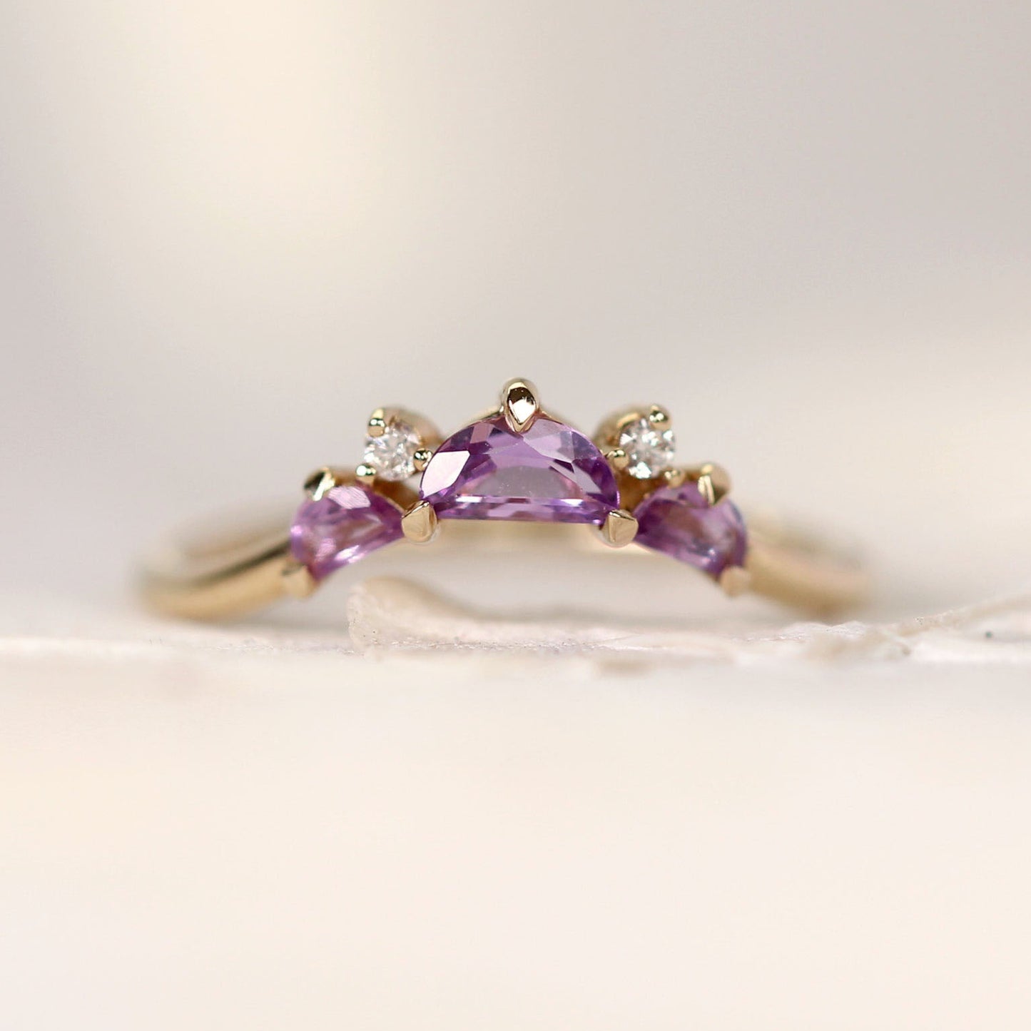 A yellow gold ring featuring a central Halfmoon cut purple gemstone flanked by two smaller marquise-cut purple gemstones and two small clear accent stones, photographed on a soft cream background.