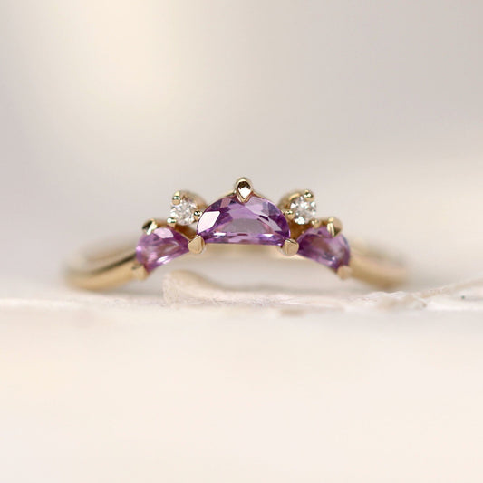 A yellow gold ring featuring a central Halfmoon cut purple gemstone flanked by two smaller marquise-cut purple gemstones and two small clear accent stones, photographed on a soft cream background.