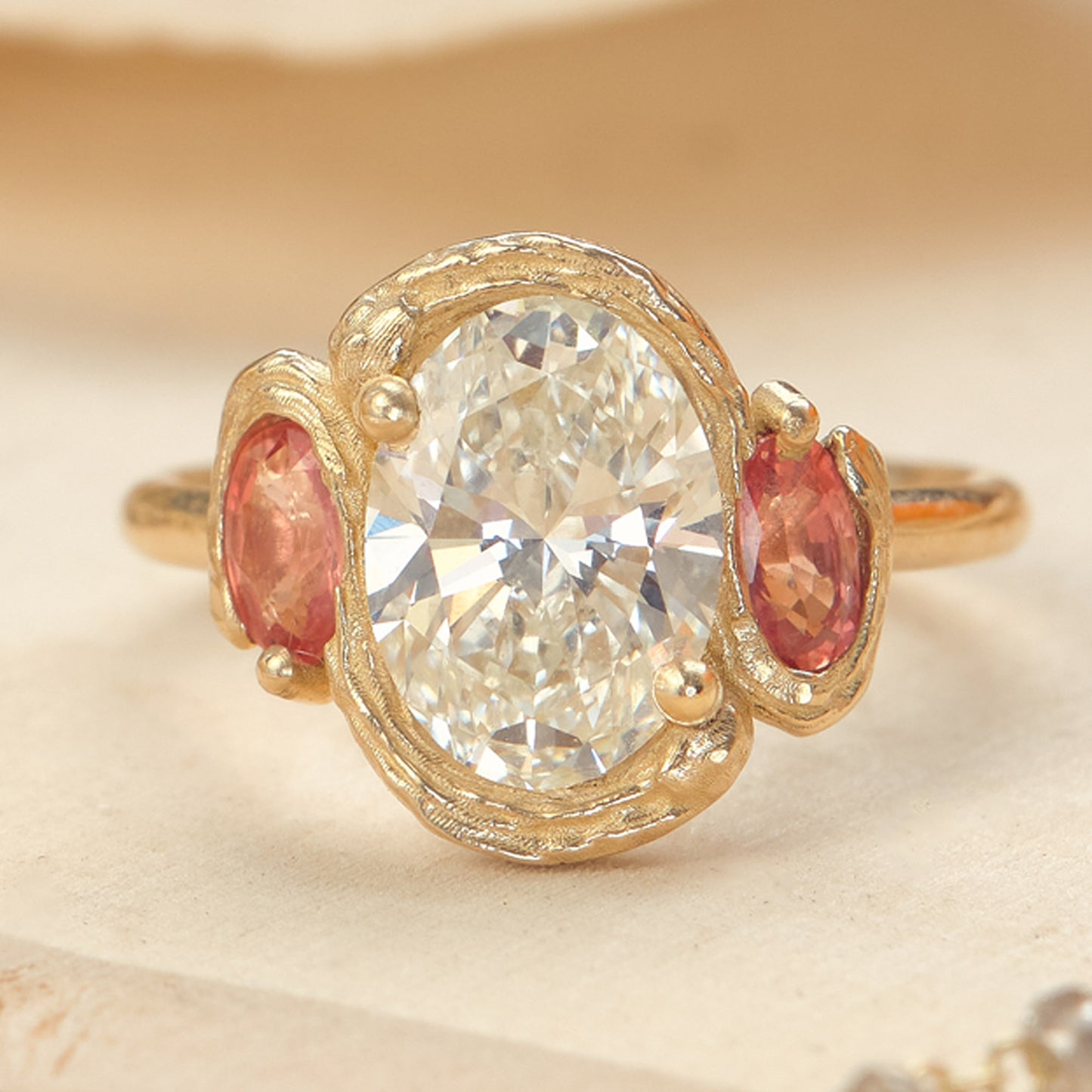A gold ring featuring a large oval clear center stone and two marquise-cut pink side stones, photographed close up on a soft beige background.